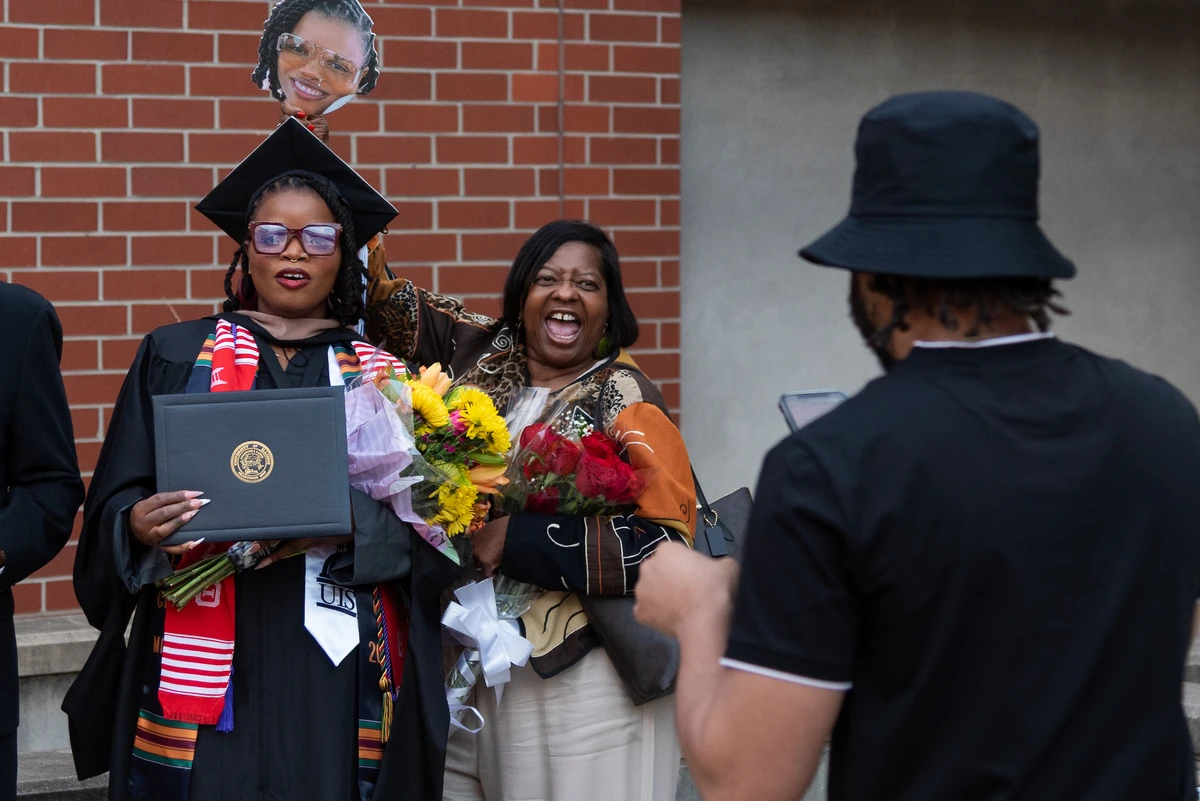 A graduate holding a diploma and bouquet poses for a photo with a beaming woman who holds a cutout of her face, while another person captures the moment on a phone.